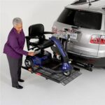 An older woman loads a mobility scooter onto a vehicle lift attached to the back of an SUV. The lift platform is extended, holding the scooter securely behind the parked car.