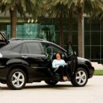 A woman sits smiling in the driver’s seat of a black SUV with the door open, parked on a paved area in front of a modern building and palm trees.