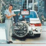 A smiling woman stands next to a car with a wheelchair lift attached to its rear, holding onto the wheelchair. The scene is outdoors in a driveway with a stone house in the background.
