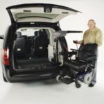 An older man stands next to a van with its rear door open, using a mechanical lift to load or unload a power wheelchair from the vehicle.