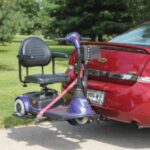 A mobility scooter attached to the back of a red car using a lift device, with a pink safety strap securing the scooter. The car is parked outdoors near grass and trees.