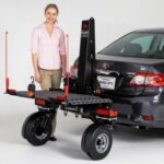 A woman stands next to a gray car with a rear-mounted platform lift attached, designed for transporting heavy equipment or mobility devices. The lift has large wheels and metal arms for support.