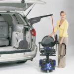 A woman uses a mechanical lift attached to a minivan to load or unload a mobility scooter. The van’s rear hatch is open, showing the lift mechanism inside.