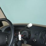 Close-up of a car dashboard with a hand control device attached to the steering column, designed to help people with physical disabilities operate the vehicle. The speedometer and air vent are also visible.