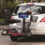 A mobility scooter is mounted on a Harmar lift attached to the back of a silver car, parked outdoors with trees in the background.