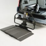 A car with its rear hatch open shows a metal platform wheelchair lift extended to the ground, ready to assist with loading or unloading a wheelchair into the vehicle.