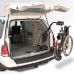 A white minivan with its rear hatch open shows a mechanical lift arm loading a folded wheelchair into the spacious cargo area.