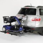 A silver SUV with a mobility scooter mounted on a rear hitch carrier; the scooter is blue and secured to a black platform attached to the vehicles back.