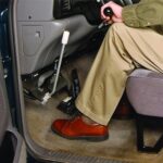 A person wearing tan pants and brown shoes sits in a vehicle driver’s seat, operating hand controls for driving, with one hand on a lever and the other on the gear shift.