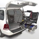 A van with its rear hatch open uses a mechanical lift to load an electric wheelchair into the back of the vehicle.
