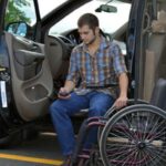 A man in a plaid shirt sits in a wheelchair next to an open car door, holding a remote control device, with the car parked in a marked parking space.