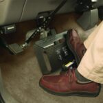 A person wearing brown shoes operates a hand control device for driving, positioned near the brake and accelerator pedals inside a car. The cars interior floor and pedal area are visible.