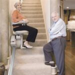 An elderly woman sits on a stair lift at the bottom of a carpeted staircase, smiling, while an elderly man stands nearby on the stairs, also smiling. The setting appears to be a home interior.