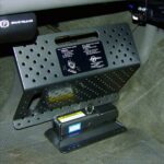 A black metal lockbox mounted under a vehicle dashboard, secured to the floor. The lockbox has ventilation holes and a handle, with a label and key lock mechanism visible on the front. Carpeted flooring surrounds the box.