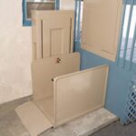 A beige vertical platform lift is installed next to a blue railing and window, providing wheelchair access between different levels of a building.