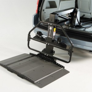 A mobility scooter or wheelchair lift is attached to the rear of a vehicle, with the platform lowered to the ground, ready for loading or unloading assistive devices.