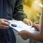 A person hands over a car key and a document on a clipboard to another person, who is signing the paperwork with a pen. The setting appears to be outdoors.