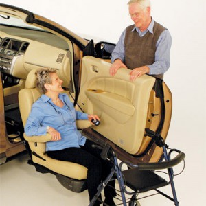 An older woman sits in a swivel car seat next to an open car door, smiling at an older man standing outside. A walker is positioned beside her, suggesting accessible vehicle features.