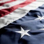 Close-up of the United States flag, showing detailed stitching on the white stars against a blue background, with part of the red and white stripes visible at the top.