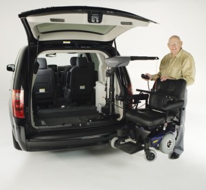 An older man stands next to a van with its rear door open, using a mechanical lift to load or unload a power wheelchair from the vehicle.