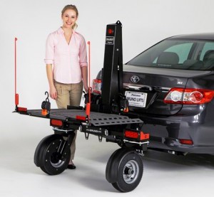 A woman stands next to a gray car with a rear-mounted platform lift attached, designed for transporting heavy equipment or mobility devices. The lift has large wheels and metal arms for support.