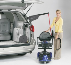 A woman uses a mechanical lift attached to a SUV to load or unload a mobility scooter from the back of the vehicle. The SUV’s trunk is open, and the woman is smiling at the camera.