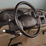 A car interior showing a steering wheel with attached hand controls for driving, designed to assist people with disabilities. The dashboard and control panel are also visible.