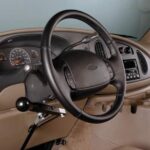 A car interior showing a steering wheel with a hand control lever installed, designed to assist drivers with limited leg mobility in operating the vehicle.