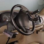 A car interior features a steering wheel equipped with hand controls designed for drivers with physical disabilities, with levers and attachments to assist with acceleration and braking.