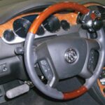 A vehicle steering wheel with a wood and gray finish, equipped with a spinner knob for easier turning, with dashboard gauges and controls visible behind it.