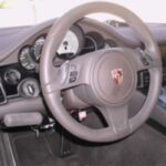 Close-up view of a Porsche car steering wheel and dashboard, featuring the Porsche logo at the center and various gauges and control buttons visible behind the wheel.