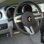 Close-up of a cars steering wheel with a Mustang emblem, dashboard gauges, and a hand control lever installed for accessibility.