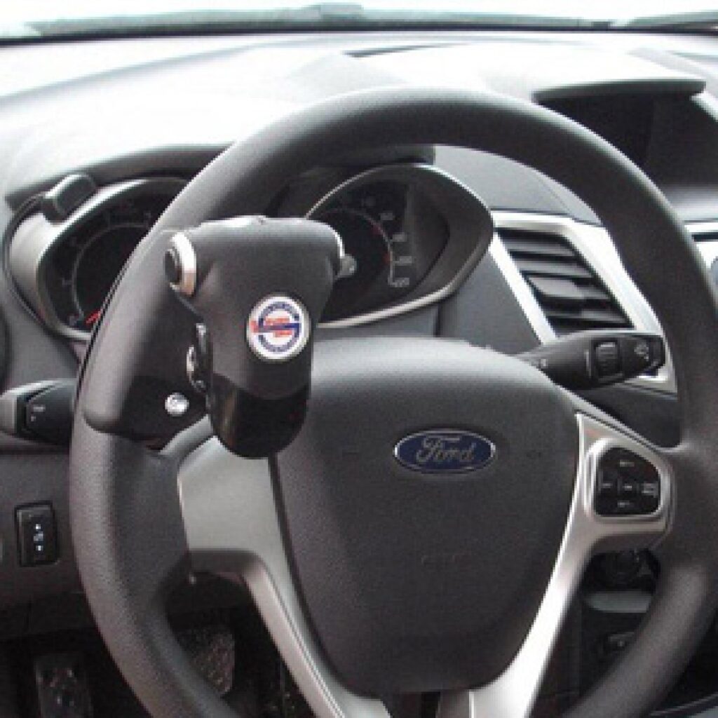Close-up of a Ford car steering wheel with a spinner knob attached, designed to help drivers turn the wheel more easily, often used for accessibility or by people with limited hand mobility. Dashboard and controls are visible.