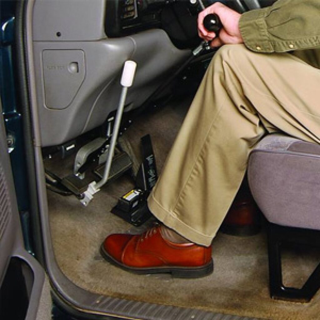 A person wearing tan pants and brown shoes sits in a vehicle driver’s seat, operating hand controls for driving, with one hand on a lever and the other on the gear shift.