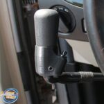 Close-up of a textured, black adaptive steering knob attached to a cars steering wheel, designed to assist drivers with limited hand mobility. A dashboard control dial is visible in the background.