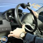A person drives a Hyundai car using a steering wheel with a spinner knob attachment. The drivers hands are positioned at 10 and 5 oclock, and they wear a wristwatch. The logo Leisure Grip appears in the corner.