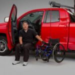 A man sits on a transfer seat extended from a red pickup truck, with a wheelchair and a mechanical lift beside him, demonstrating vehicle accessibility features.