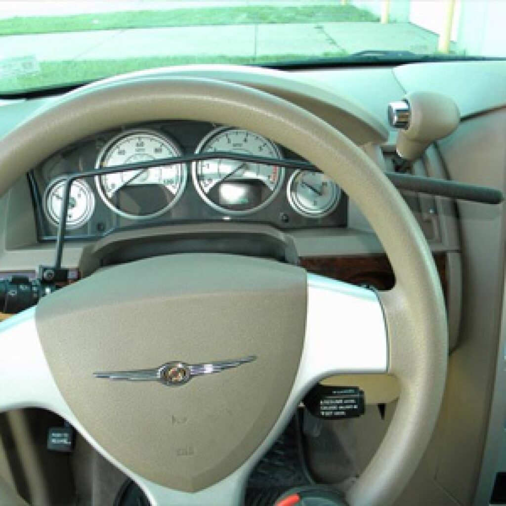 A car steering wheel with hand controls installed for accessibility, showing dashboard gauges and a beige interior.