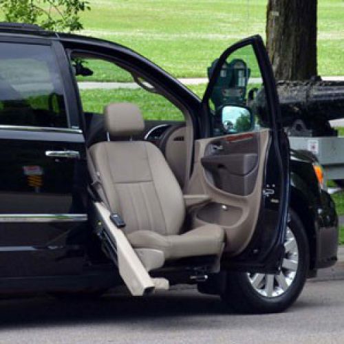 A black minivan with its front passenger door open, showing a beige swivel seat extended outward, designed for easier access or transfer for people with mobility challenges.