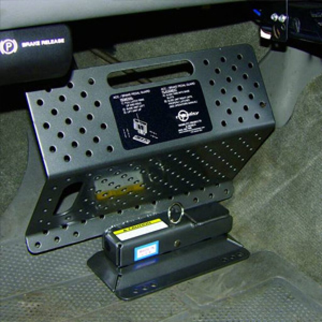 A black metal lockbox mounted under a vehicle dashboard, secured to the floor. The lockbox has ventilation holes and a handle, with a label and key lock mechanism visible on the front. Carpeted flooring surrounds the box.