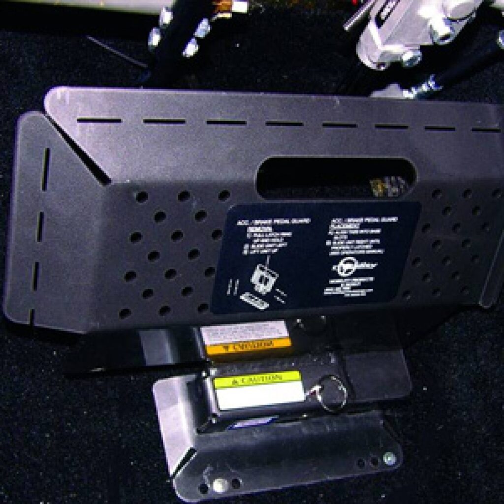 Close-up of a black metal pedal guard with ventilation holes and caution labels, installed on a vehicle floor, designed to cover and protect the pedals below.