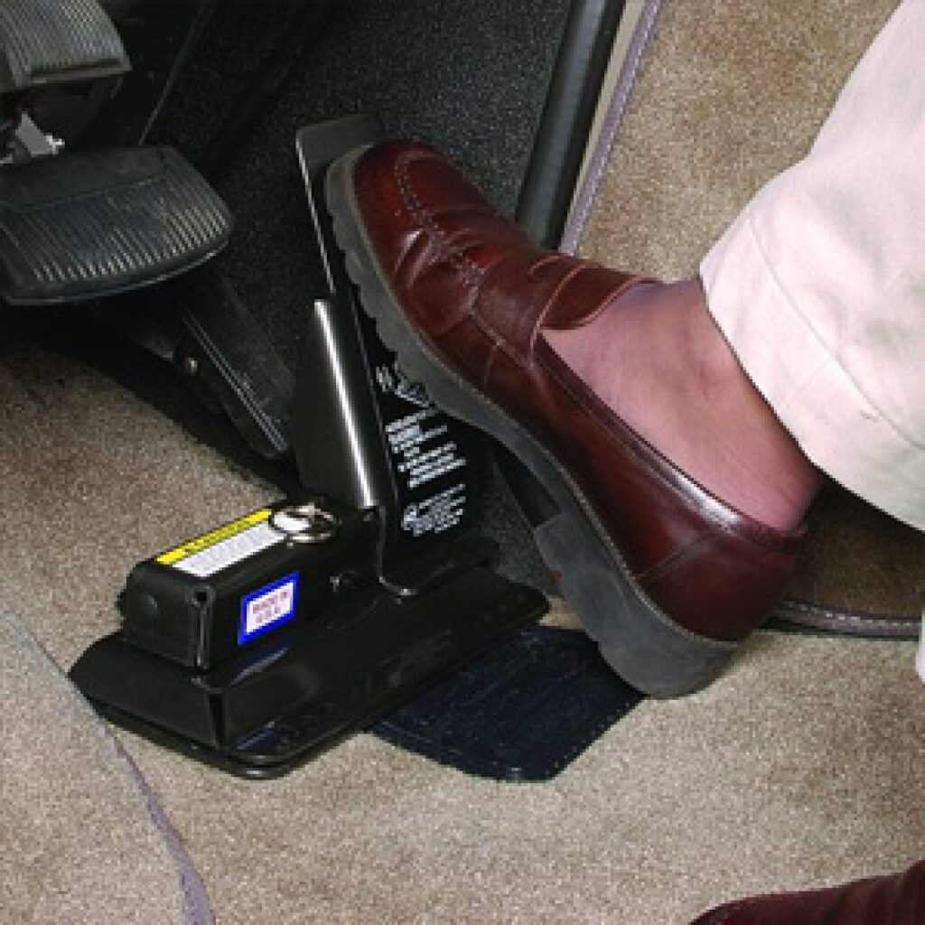 A person wearing brown dress shoes and light pants presses their foot on an emergency brake pedal inside a car with beige carpeting.