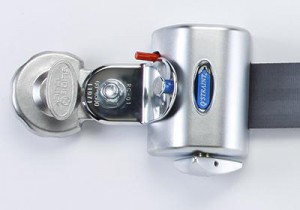 A close-up of a silver vehicle seat belt lock with a key inserted, a blue label reading OPTILOCK, and a black seat belt threaded through the device.