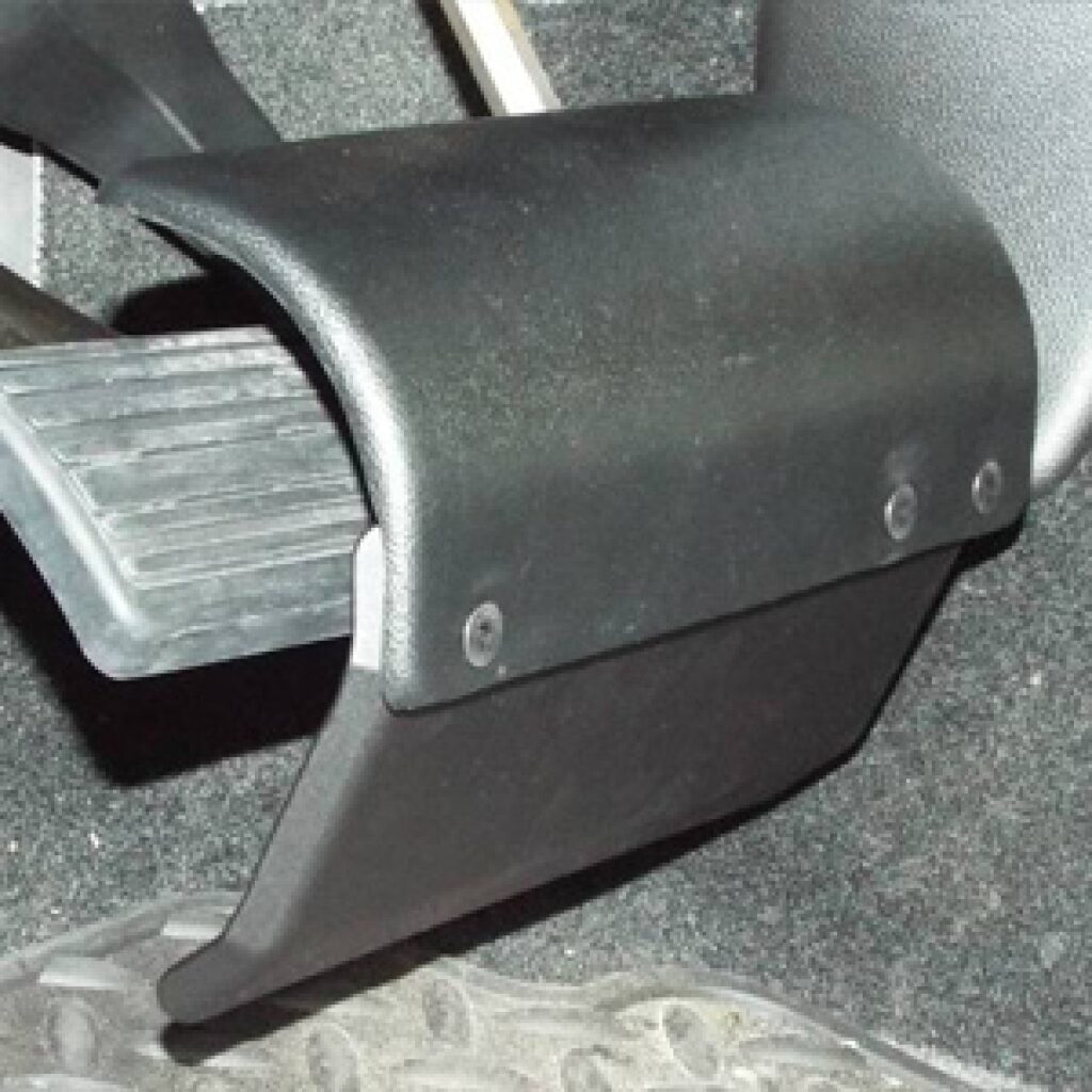 A close-up view of a car pedal with a black pedal guard installed, partially covering the pedal. The car’s carpeted floor and part of a rubber floor mat are visible underneath.