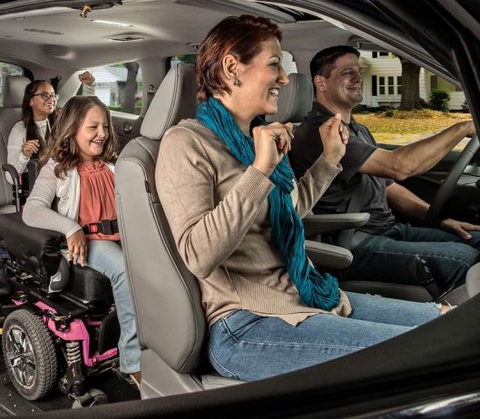 A family of four, including a child in a wheelchair, smiles and dances inside a car. The driver and passengers appear happy and engaged, enjoying a fun moment together.